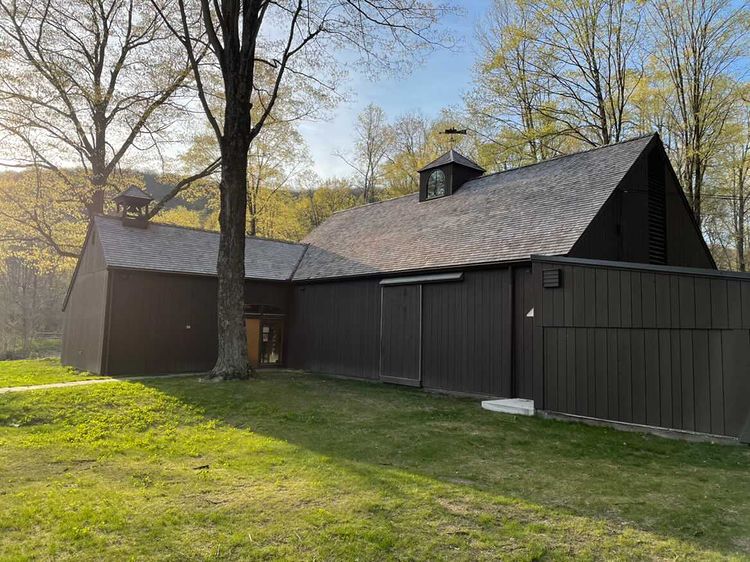 Exterior of a dark brown barn-style building surrounded by green grass and trees in early spring