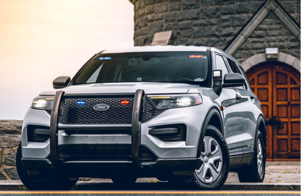 A gray Ford Explorer Police Interceptor Utility vehicle is parked in front of a stone building with a large wooden door. The vehicle has blue and red lights activated on its roof and grill. A cloudy sky provides the backdrop.