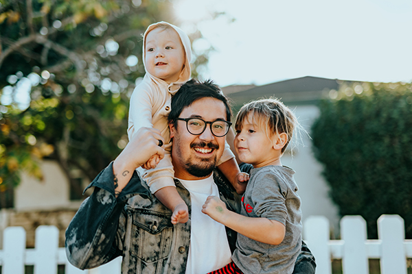 Family outside of home with white picket fence in the background. Fathering holding his two children on lap and in in hand.