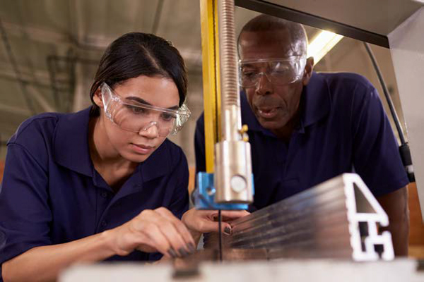 Young woman working on steel manufacturing facility with black supervisor hovering over her for direction.