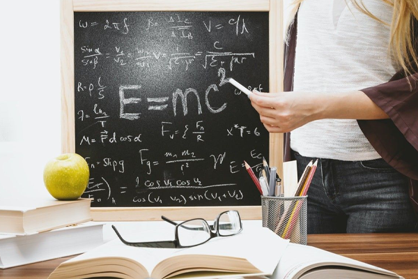 teacher standing at desk with an apple and a chalkboard in the background